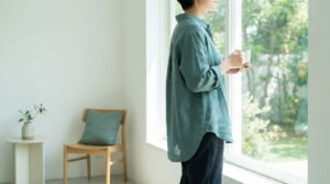 Une personne vêtue d'une chemise vert sauge regarde par la fenêtre en tenant une tasse, dans un intérieur minimaliste.