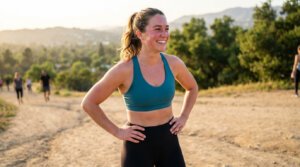Jeune femme souriante en tenue de sport, visage rougi par l'effort, sur un sentier de terre avec des personnes en arrière-plan.